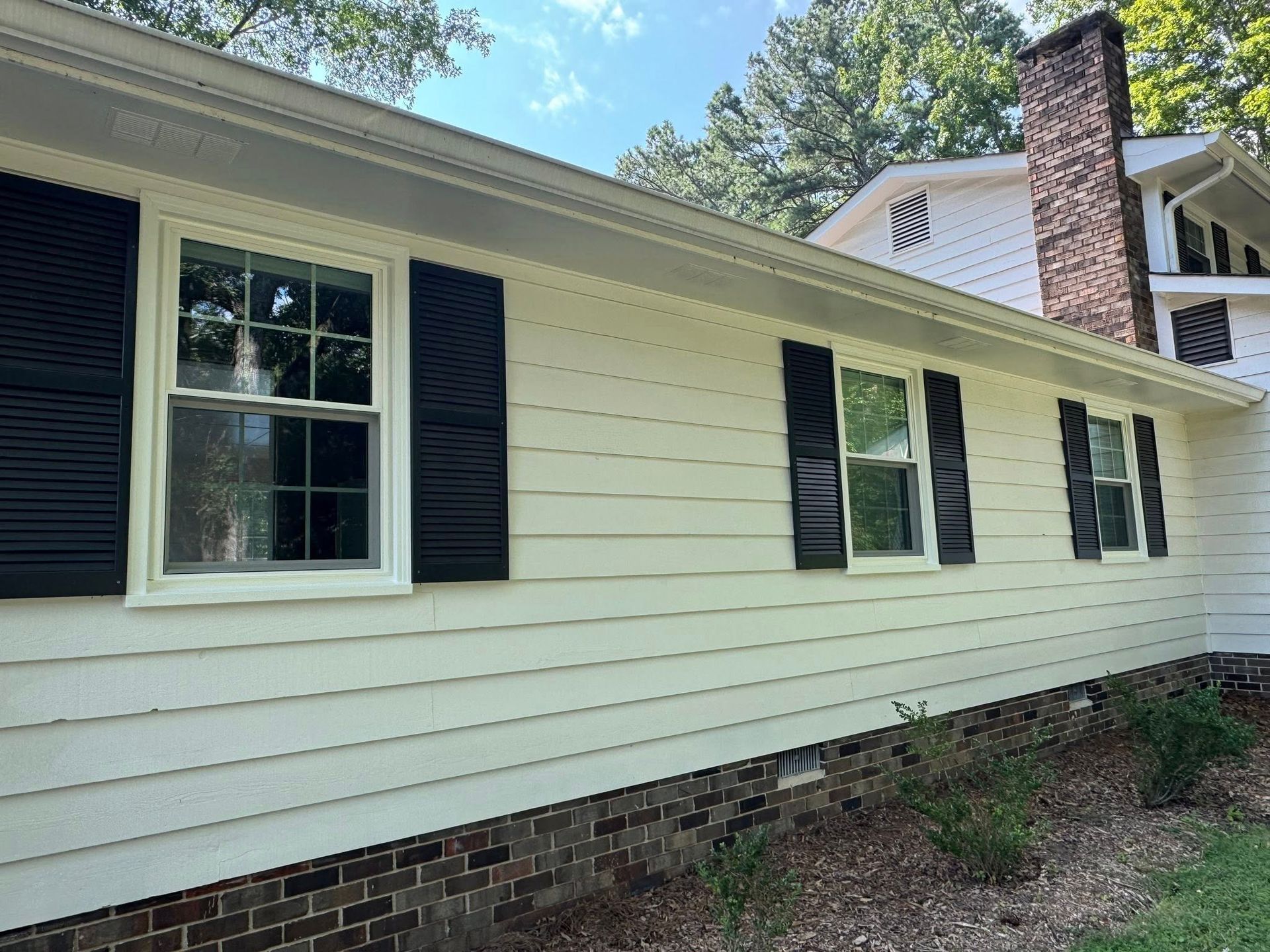 White house with black shutters and brick base; blue sky.