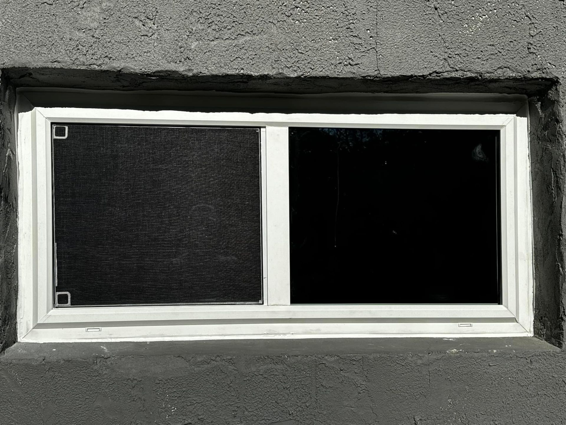 White-framed window in a gray concrete wall, with one dark, screened pane and one dark, clear pane.