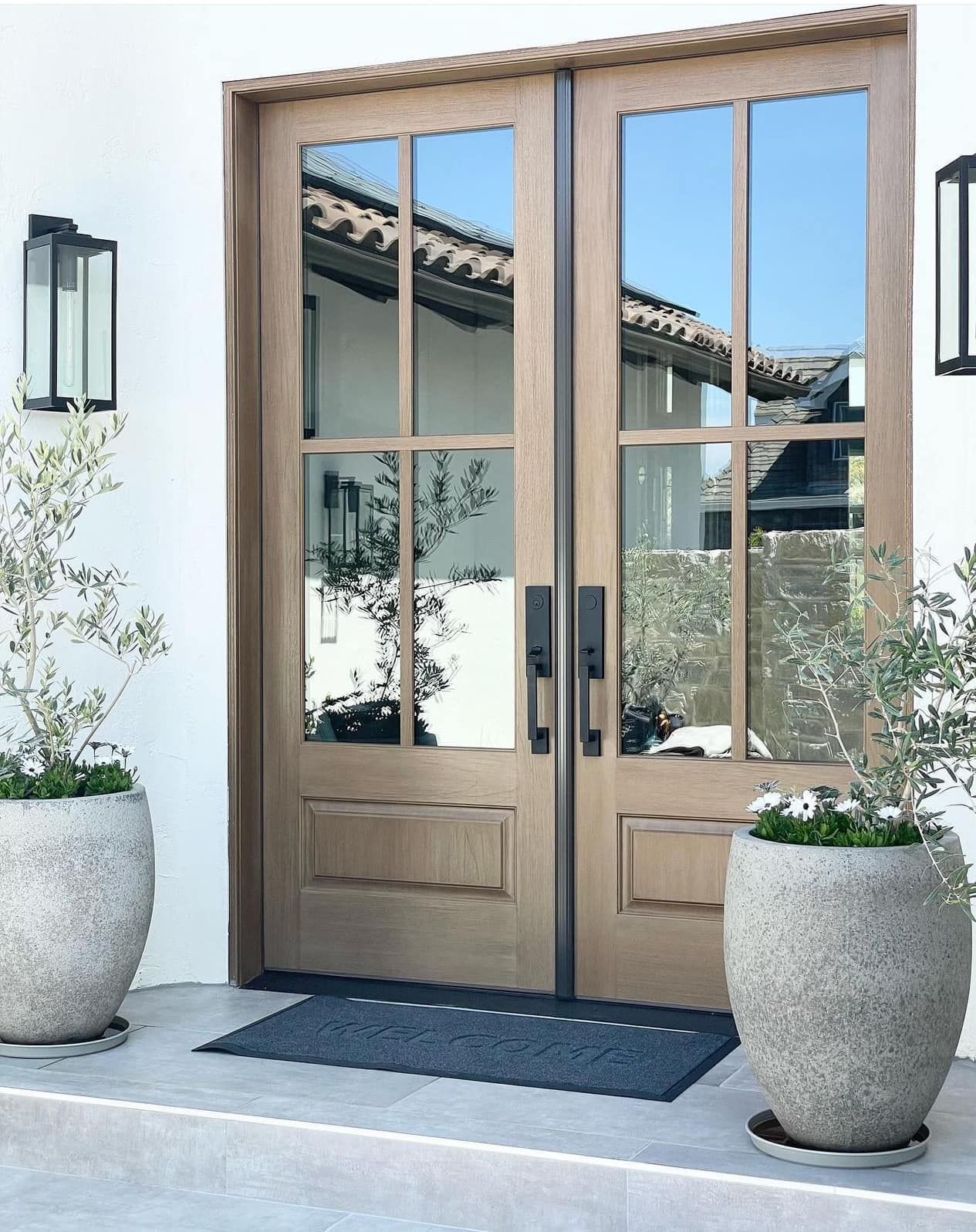 Double wooden entry doors with glass panes, flanked by potted plants and wall sconces.