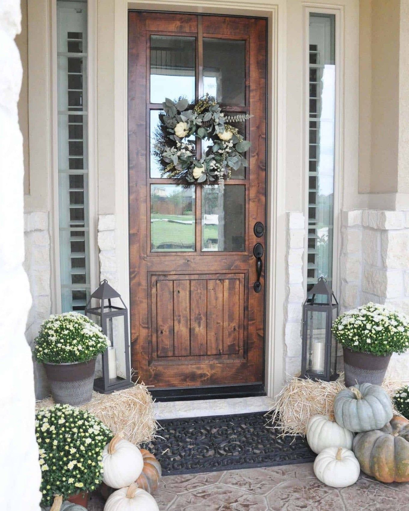 Wooden door decorated for fall with wreath, mums, pumpkins, hay bales, and lanterns.