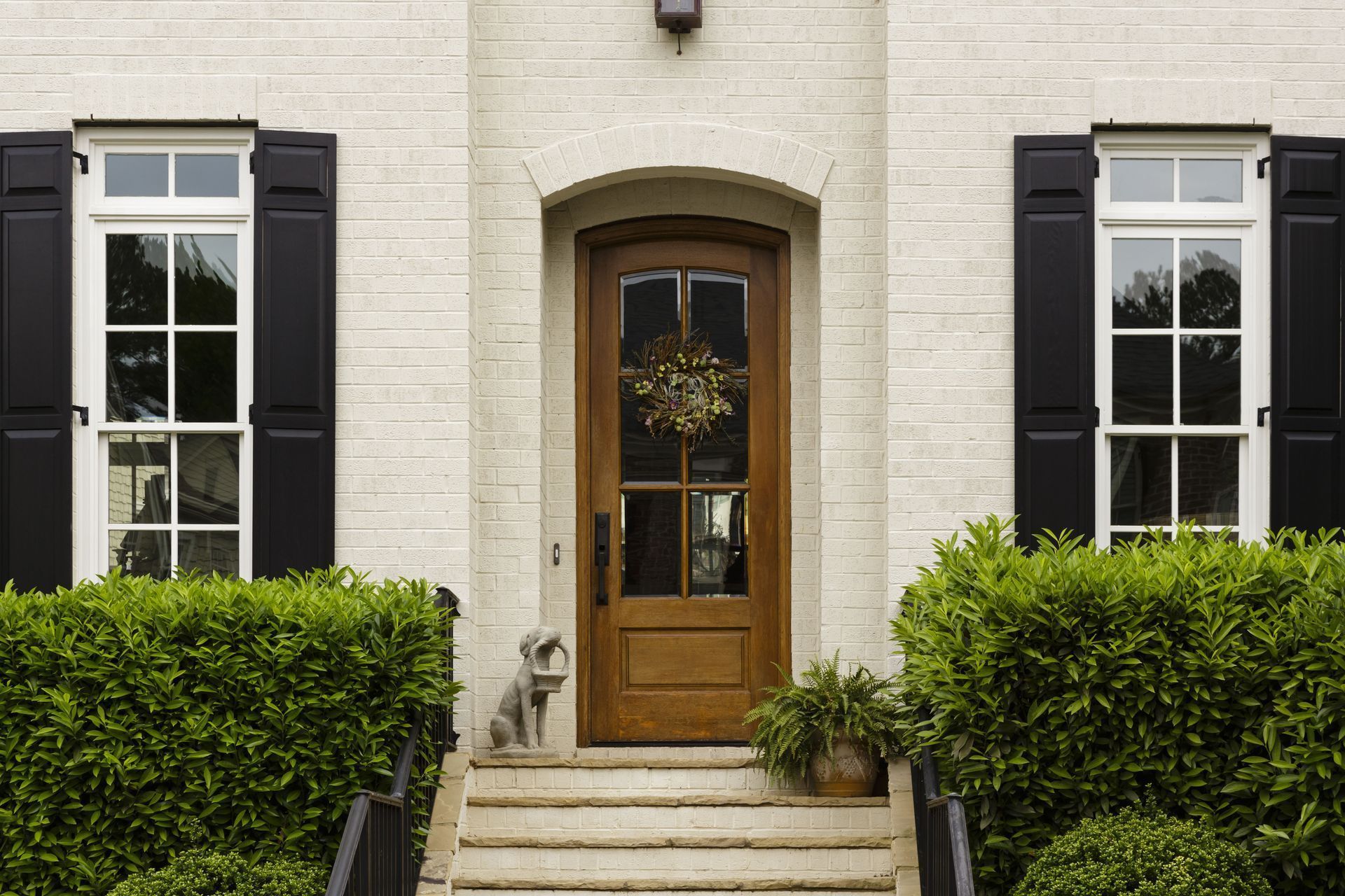 Tan brick facade with black shutters, wooden door, and manicured greenery.