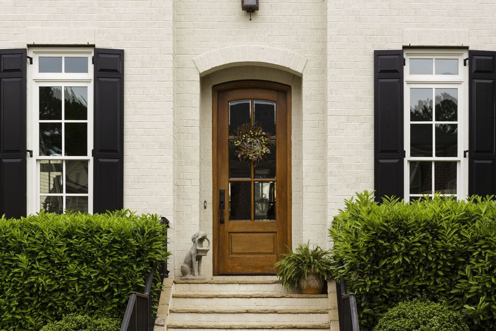 Beige brick facade with dark shutters, brown door, and green hedges.
