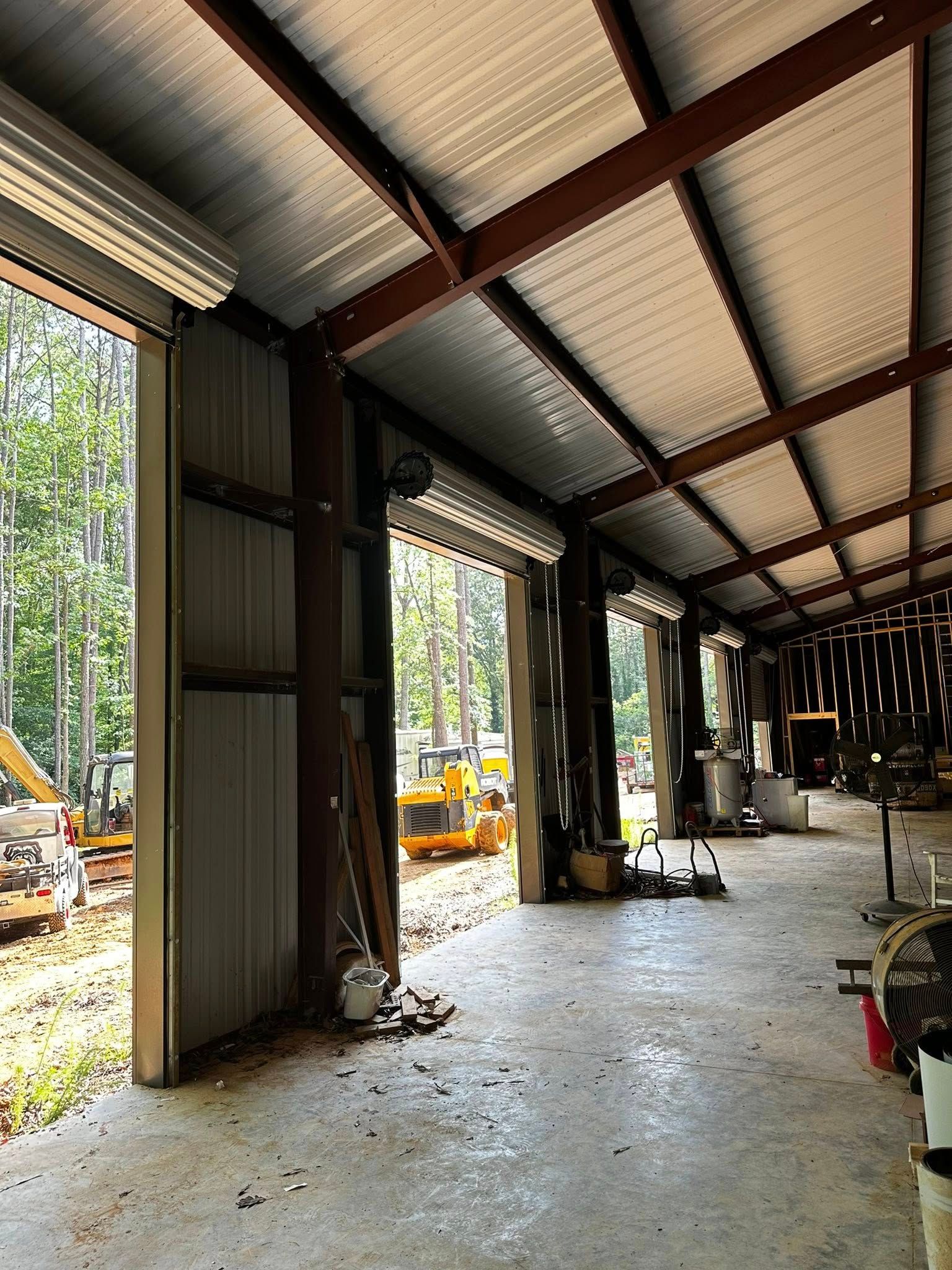 Interior of a metal building with open garage doors. Construction equipment visible outside.