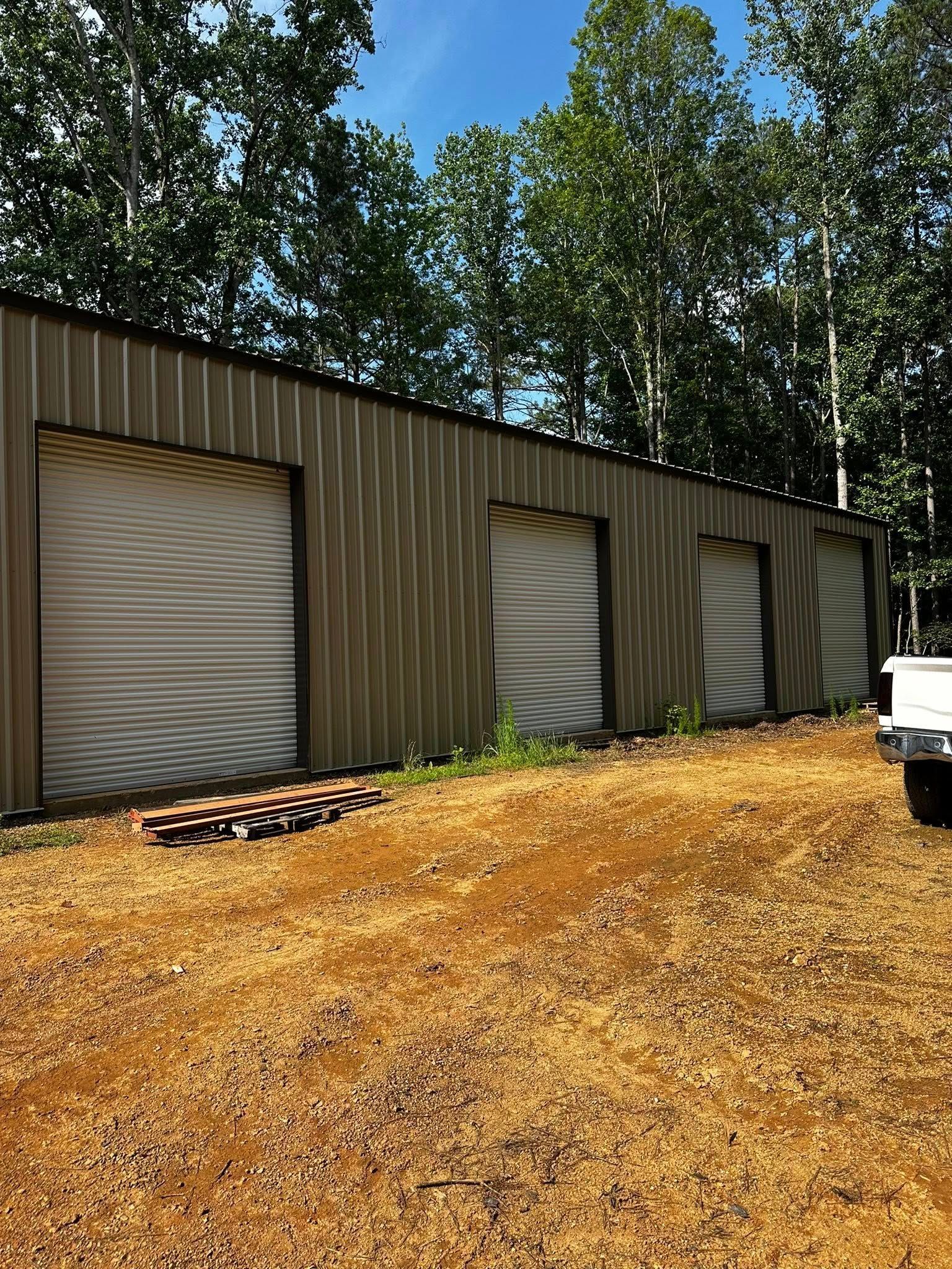 Metal garage with three roll-up doors. Gravel driveway, trees in background, sunny day.