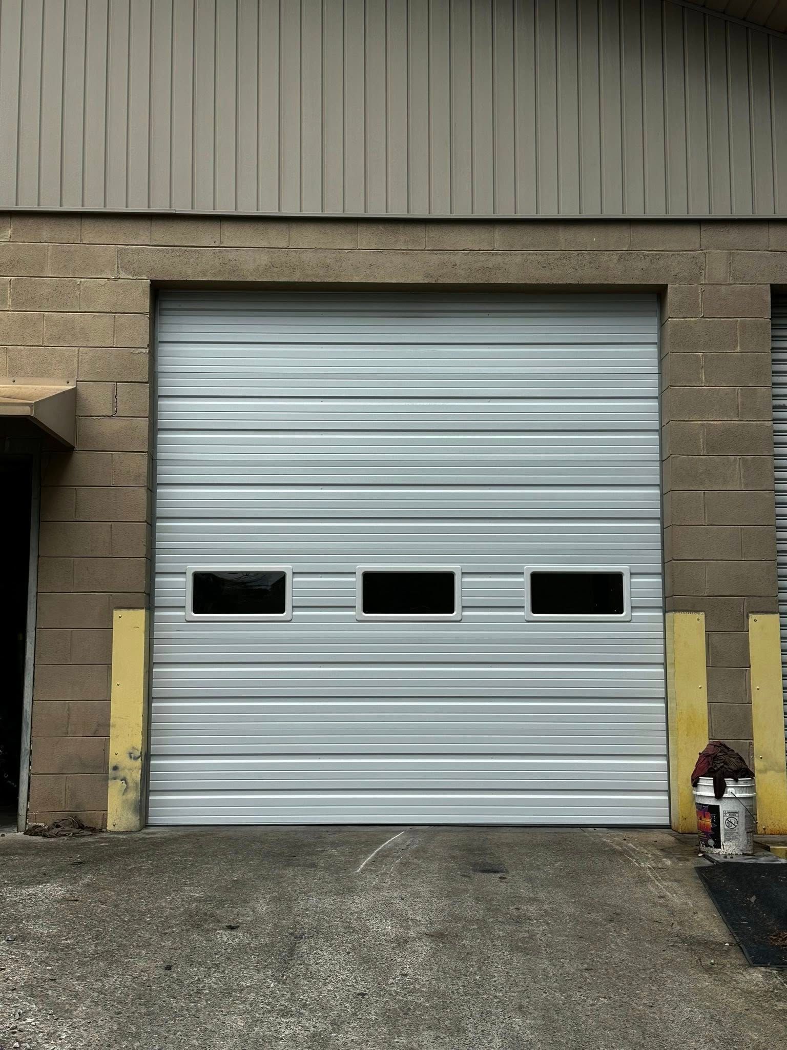 Gray garage door with three small windows. Brown brick and tan metal siding. Gravel ground.