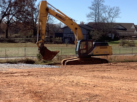 Yellow Kobelco excavator digging on a dirt lot. In the background are trees and a fence.