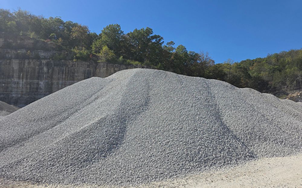 Pile of gray gravel against a hillside with trees under a blue sky.