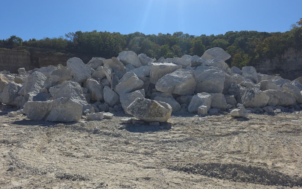 Pile of large, light-colored rocks on a light brown, dusty ground. Trees and blue sky are in the background.