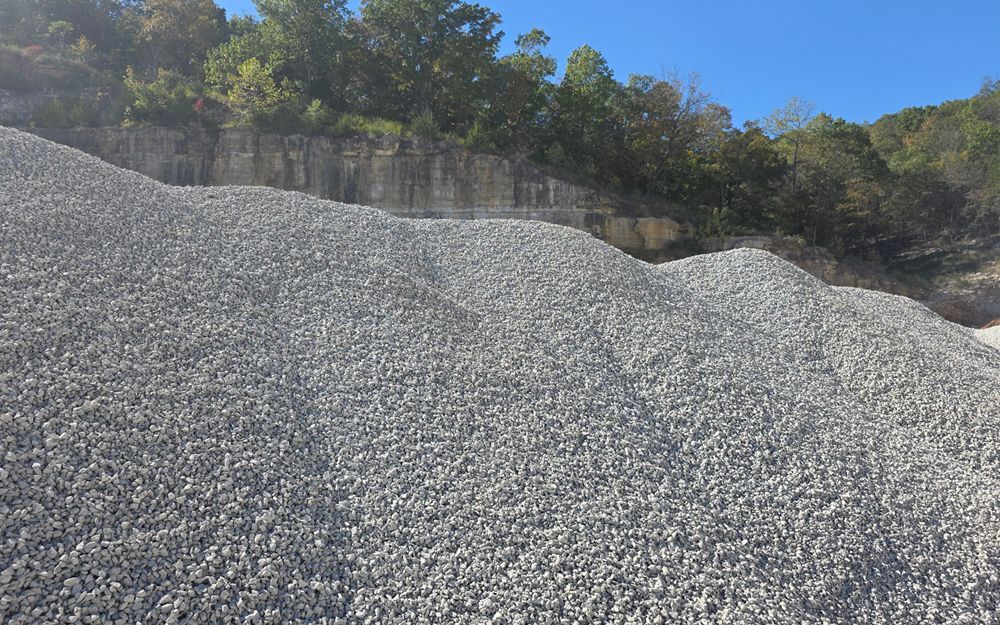 Large pile of gray gravel in front of a rock face with trees and a clear blue sky.