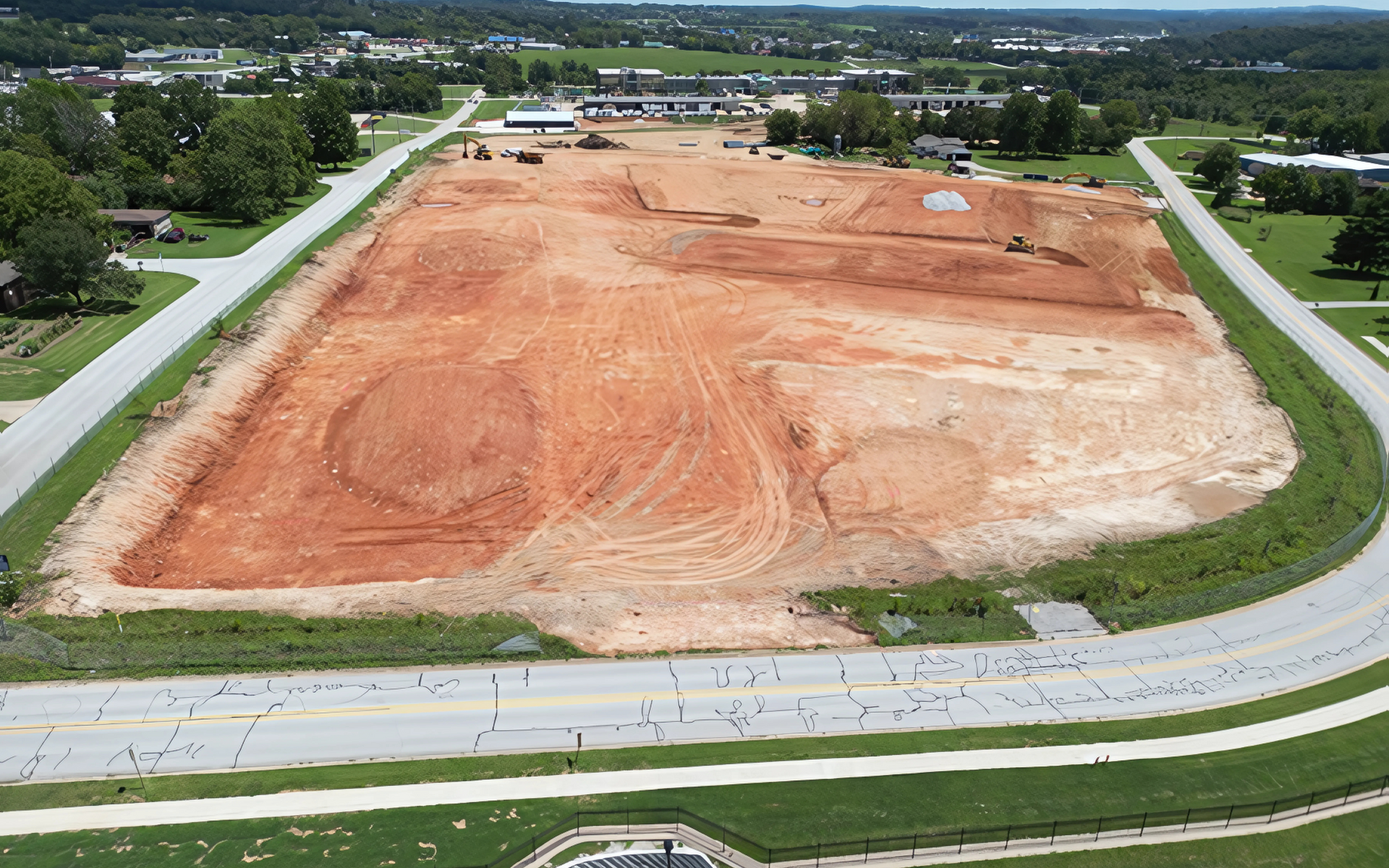 Aerial view of a large construction site with red soil surrounded by roads and grass.