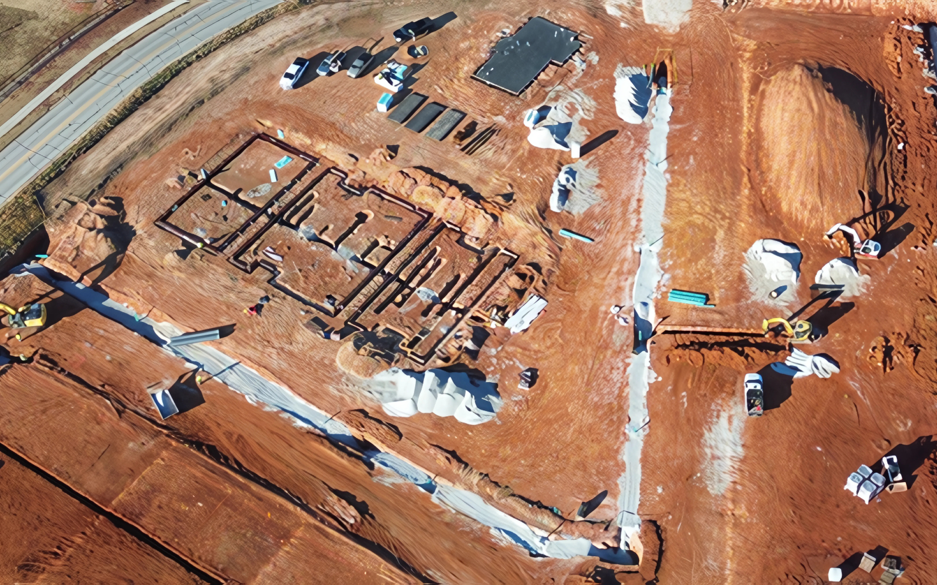 Aerial view of construction site with foundation outlines, earthmoving equipment, and dirt mounds.