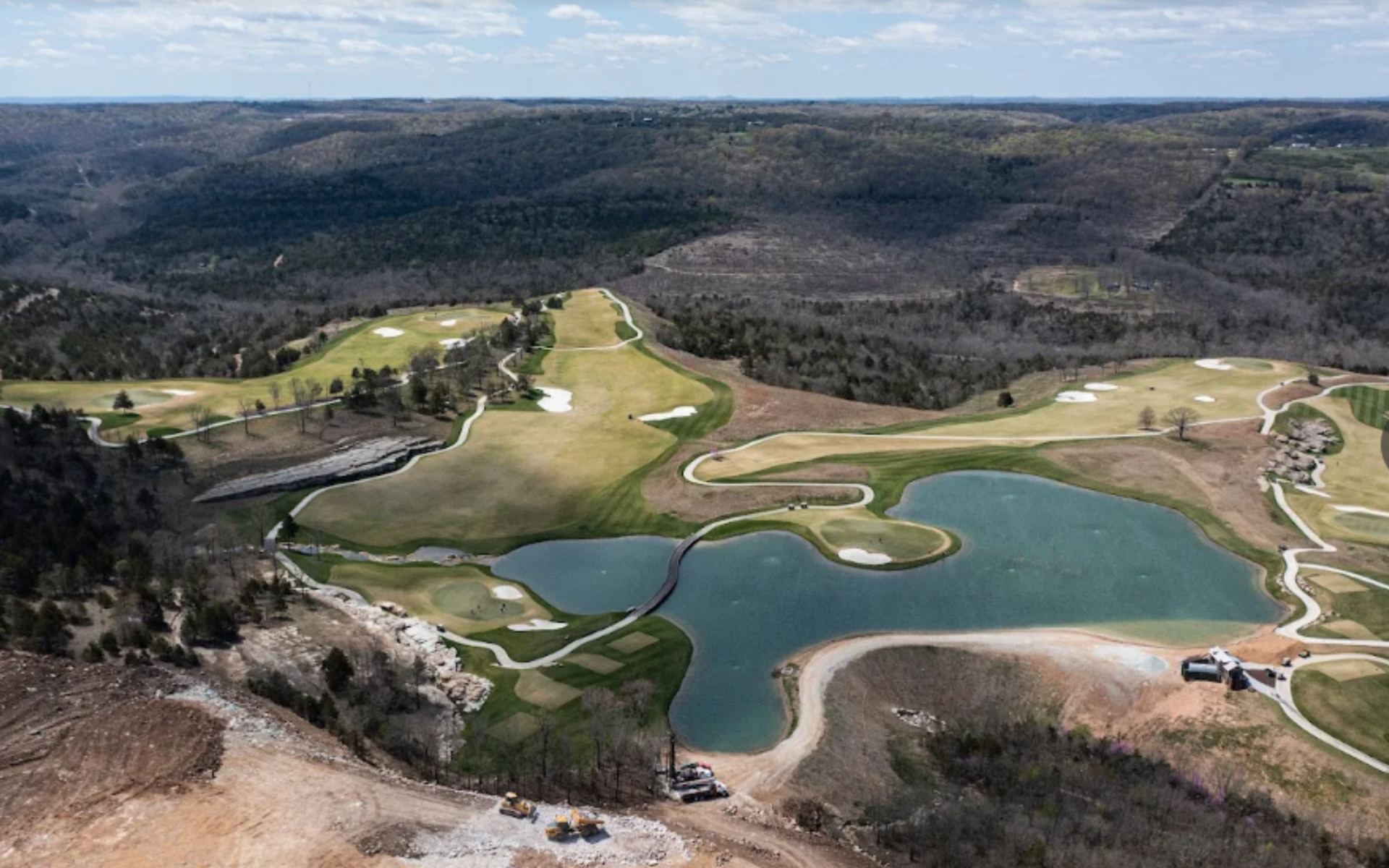 Aerial view of a golf course with a large lake, green fairways, and surrounding forest.