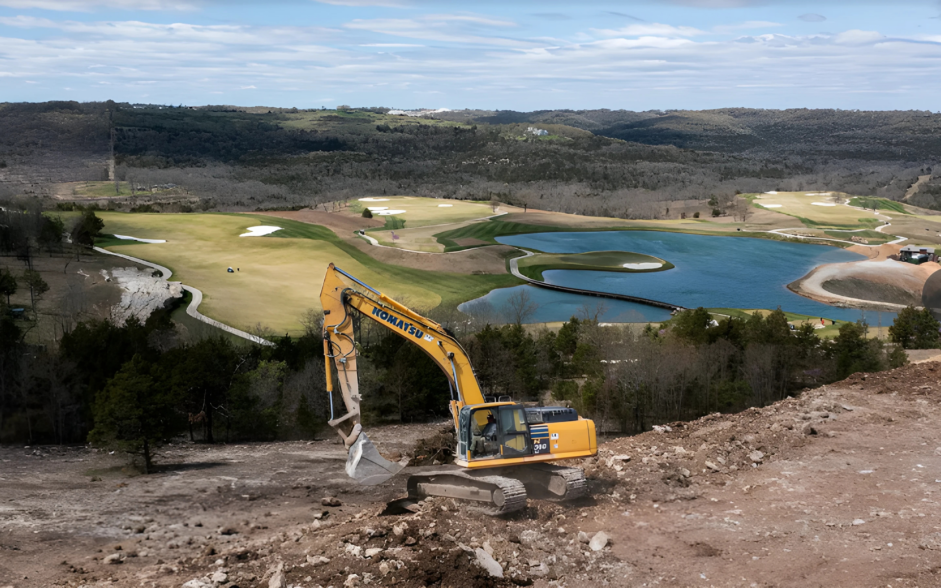 Excavator digging earth on hillside with golf course, lake, and trees in the background.