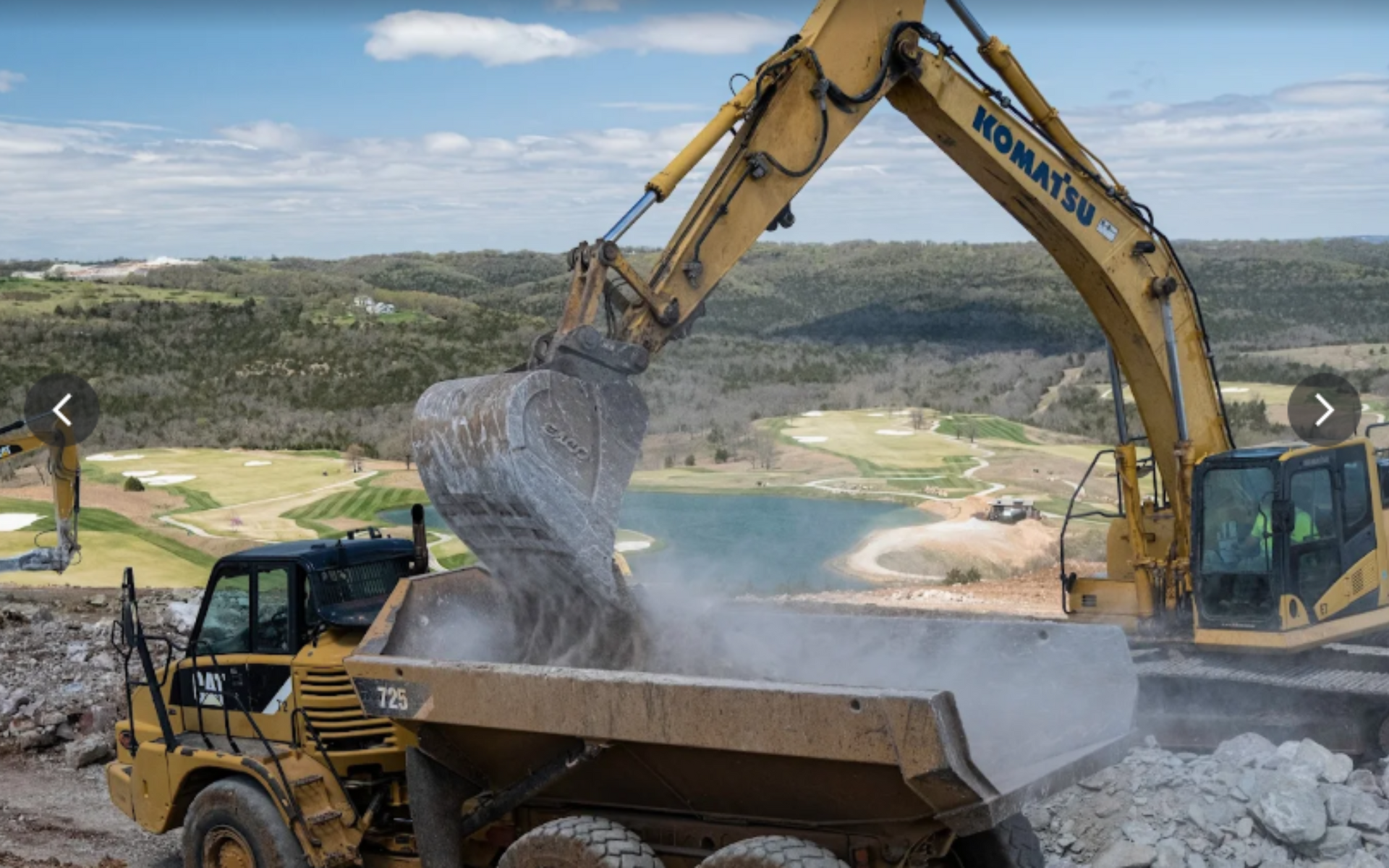 Yellow excavator loading rock into a dump truck at a construction site overlooking a lake and green landscape.