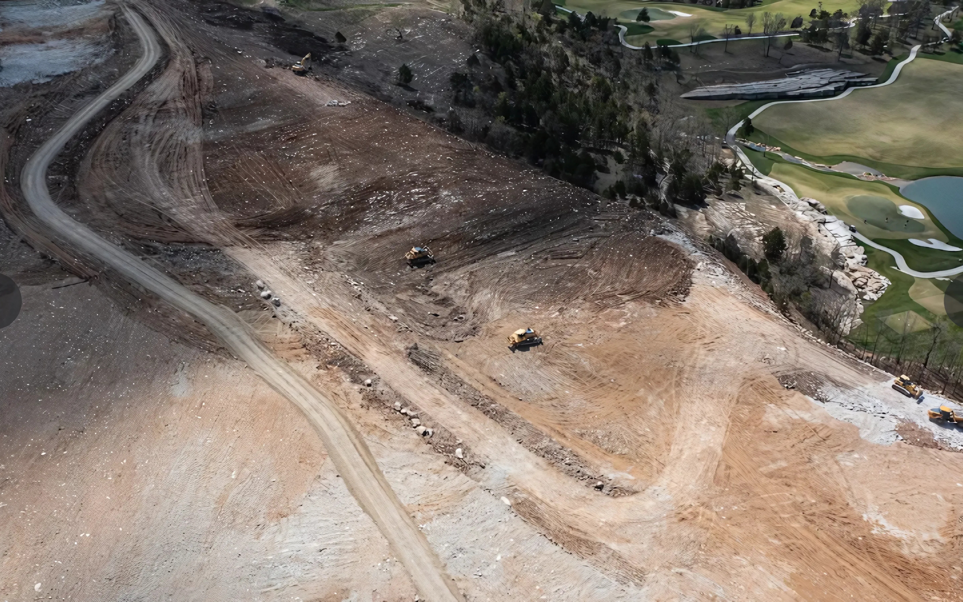 Construction site with earth-moving equipment on a hillside; a golf course and water visible nearby.