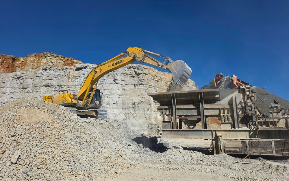 Yellow excavator loading rock into a processing machine at a quarry under a blue sky.