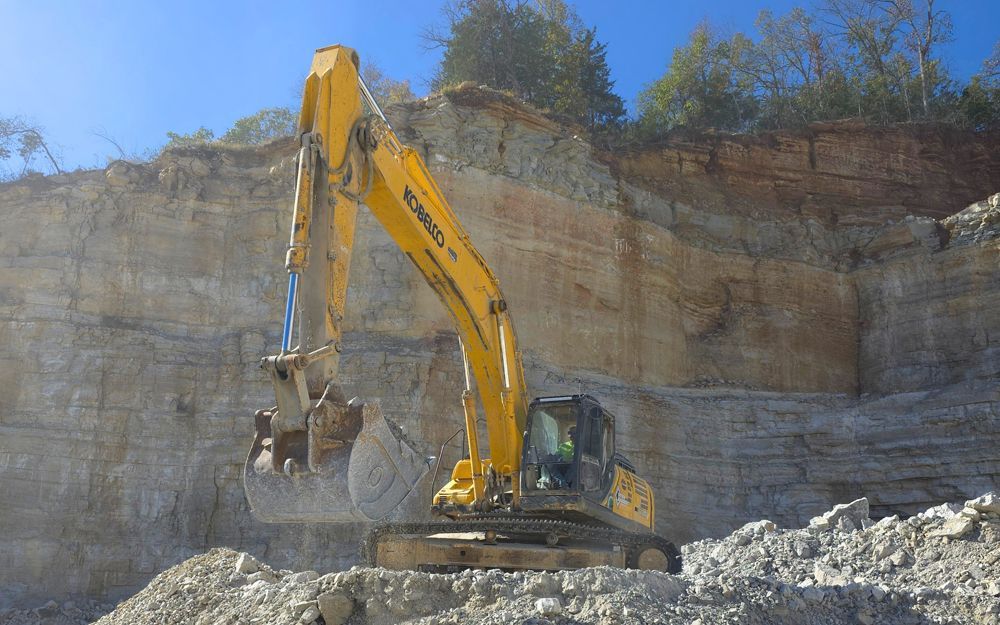 Yellow excavator digging into a limestone cliff face at a quarry on a sunny day.