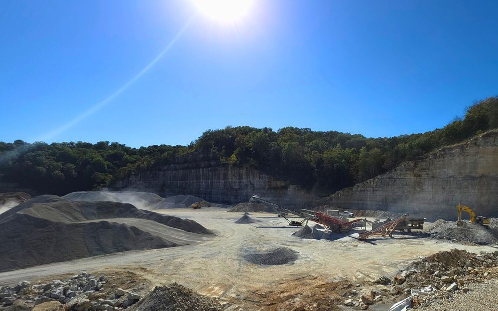 Sunny quarry with machinery and piles of gravel, against a backdrop of trees and a blue sky.