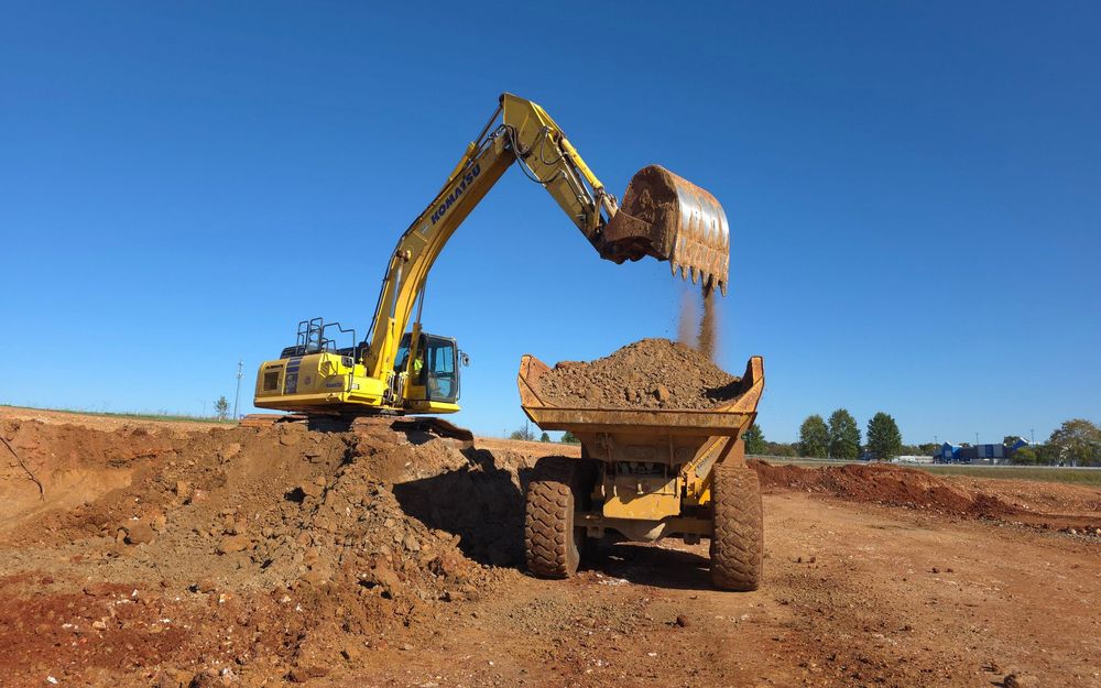 Excavator dumping dirt into a dump truck at a construction site under a clear blue sky