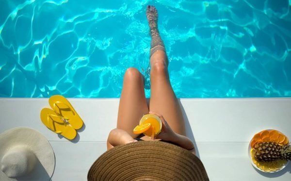 A woman is sitting on the edge of a swimming pool drinking a drink.
