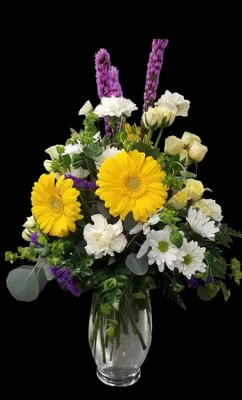 Yellow Gerber daisies, white daisies and carnations in a clear vase against a black background.
