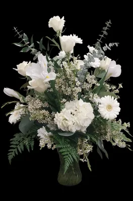 White floral arrangement in a clear vase, featuring various blooms and green foliage against a black backdrop.