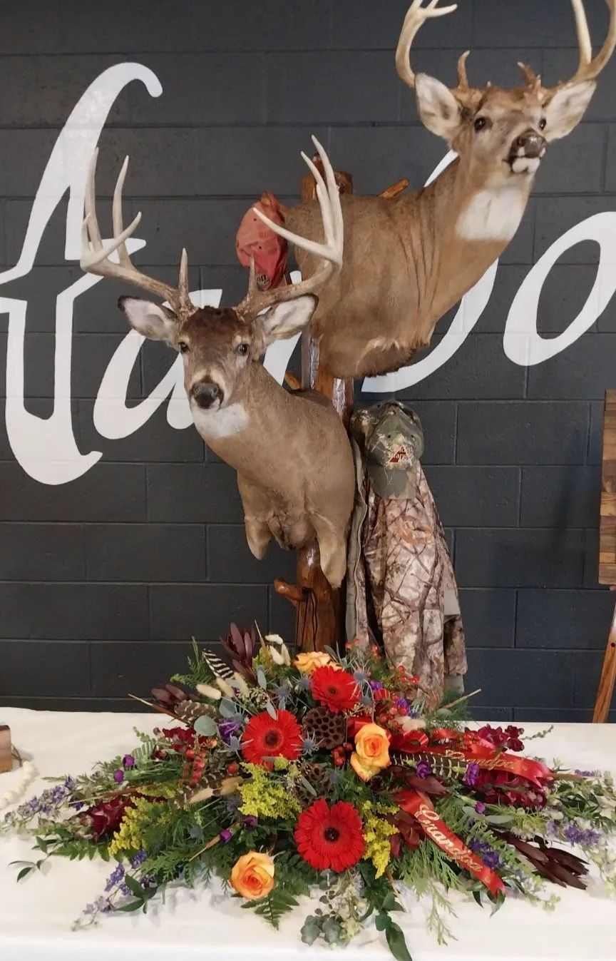 Two deer taxidermy mounts above a flower arrangement on a white table. A camo jacket is draped near the base.