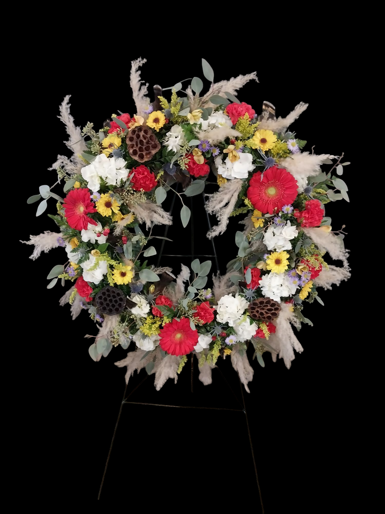 Floral wreath on a stand; white, red, and yellow flowers with dried seed pods and feathery grasses.