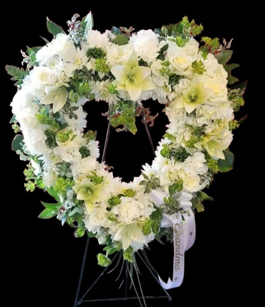 Heart-shaped floral arrangement of white flowers and greenery on a stand; a ribbon reads 
