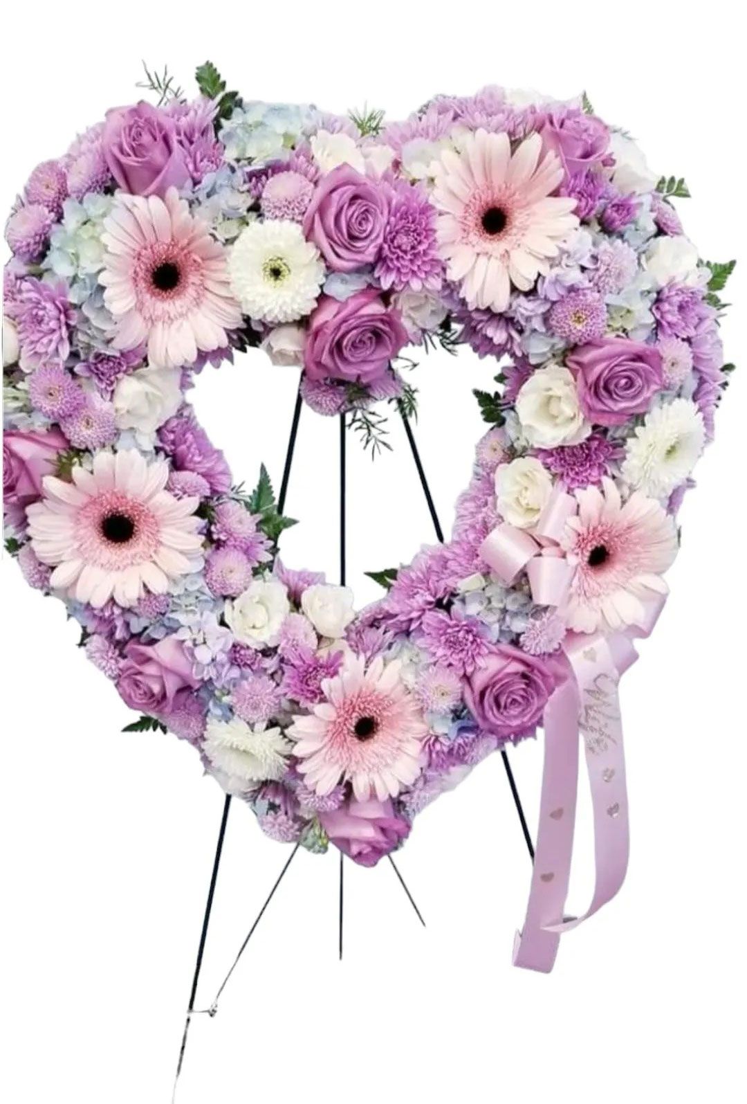Heart-shaped floral arrangement in shades of purple, pink, and white on a black easel. A light pink ribbon hangs down.