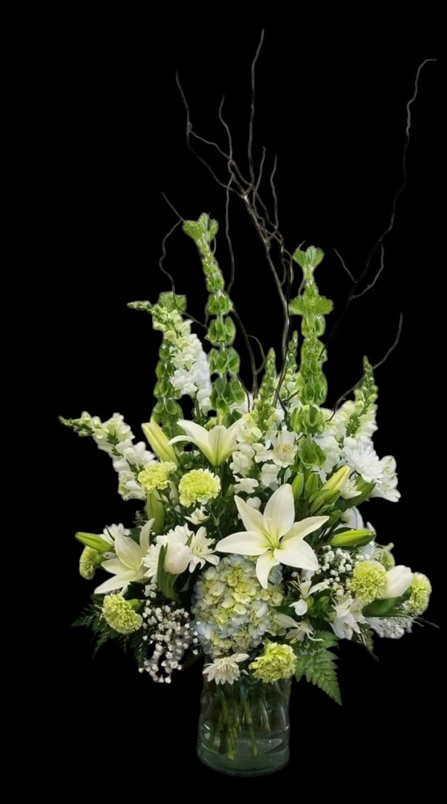 Floral arrangement of white lilies, snapdragons, and greenery in a glass vase against a black backdrop.