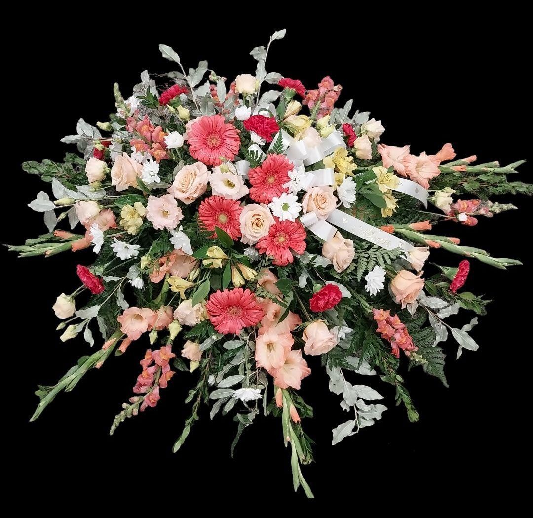 Funeral flower arrangement with pink, red, and peach blooms, set against dark background.