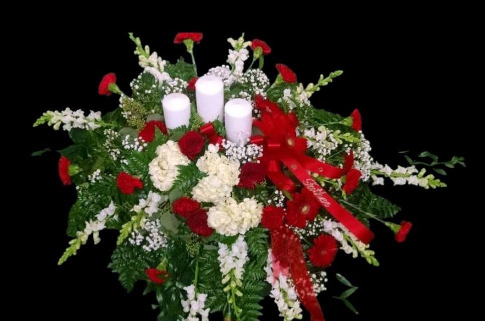 Floral arrangement with red carnations, white flowers, greenery, three candles, and a red ribbon.