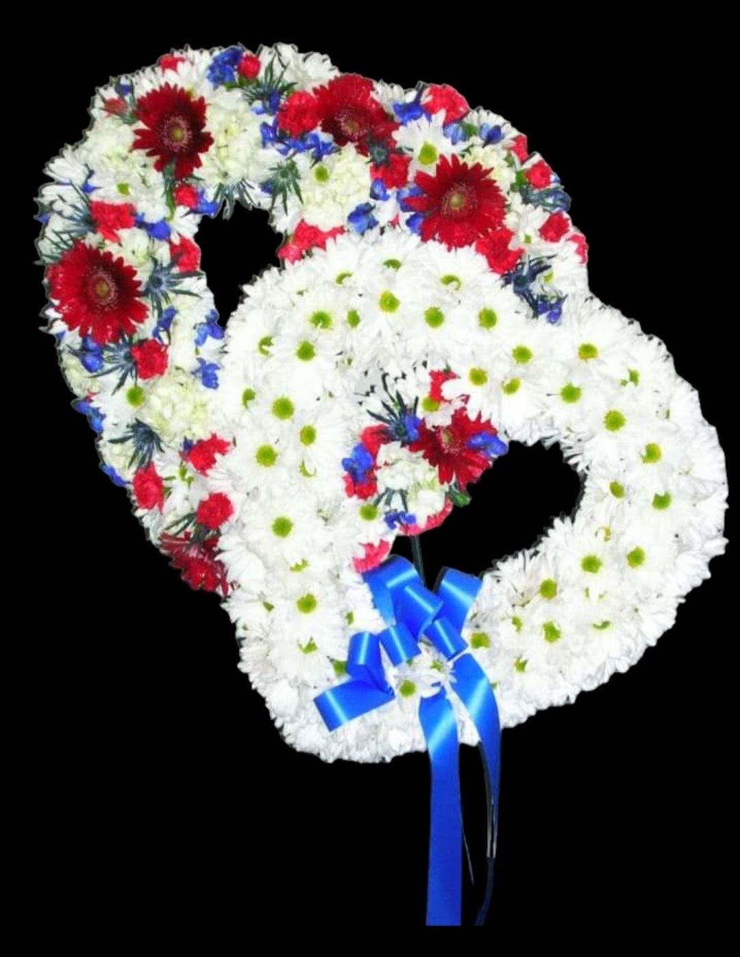 Two intertwined heart-shaped floral wreaths; one red, white, and blue, the other white. Tied with a blue ribbon.