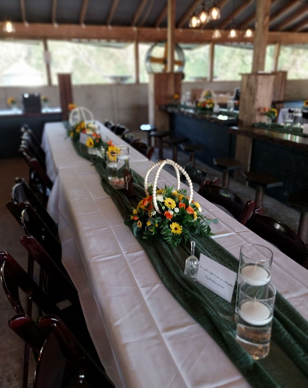 Long table set for an event, decorated with flowers and green runner, in a rustic venue with bar.