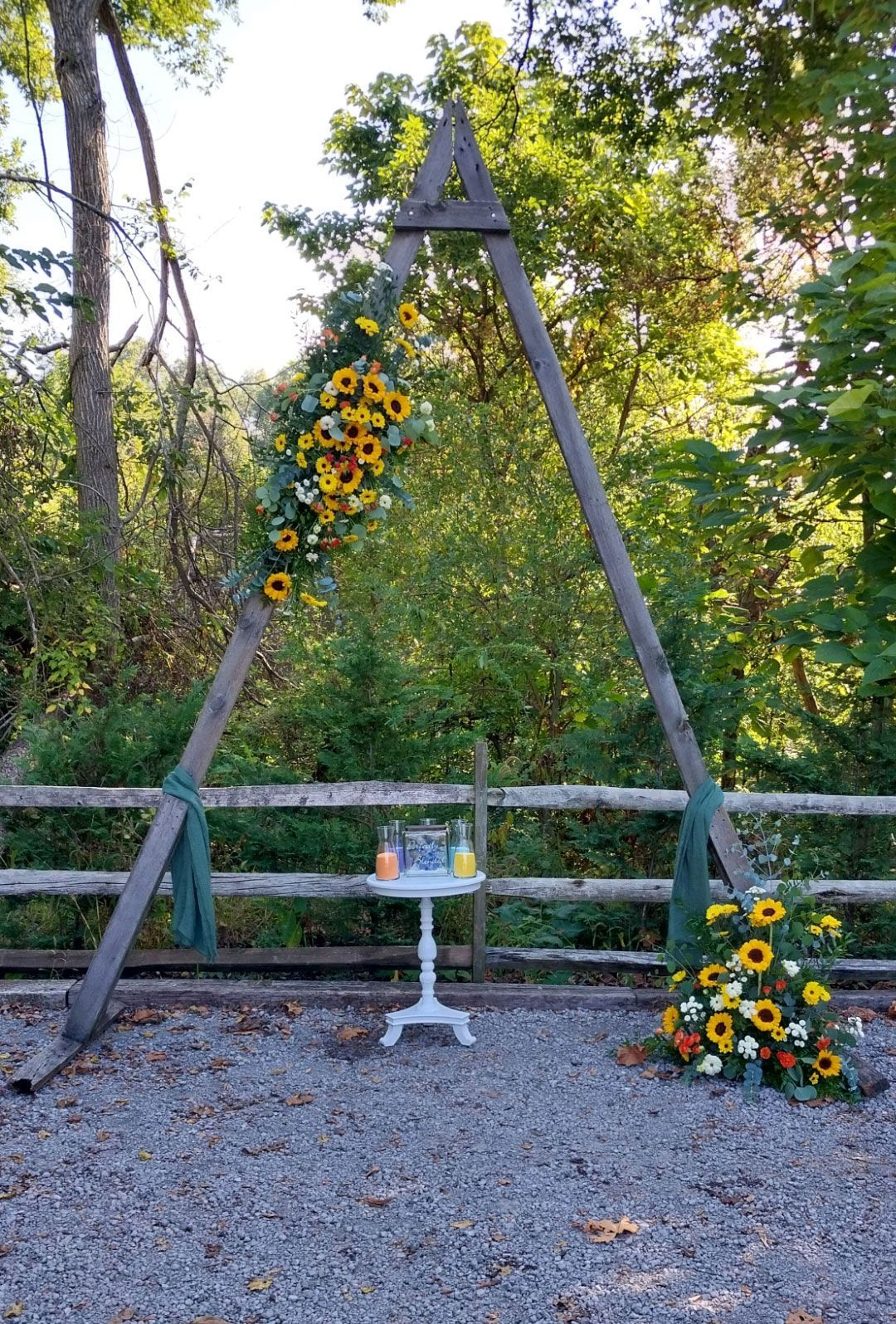 Wooden wedding arch decorated with sunflowers. Small table with sand ceremony setup, and a floral arrangement on the ground.