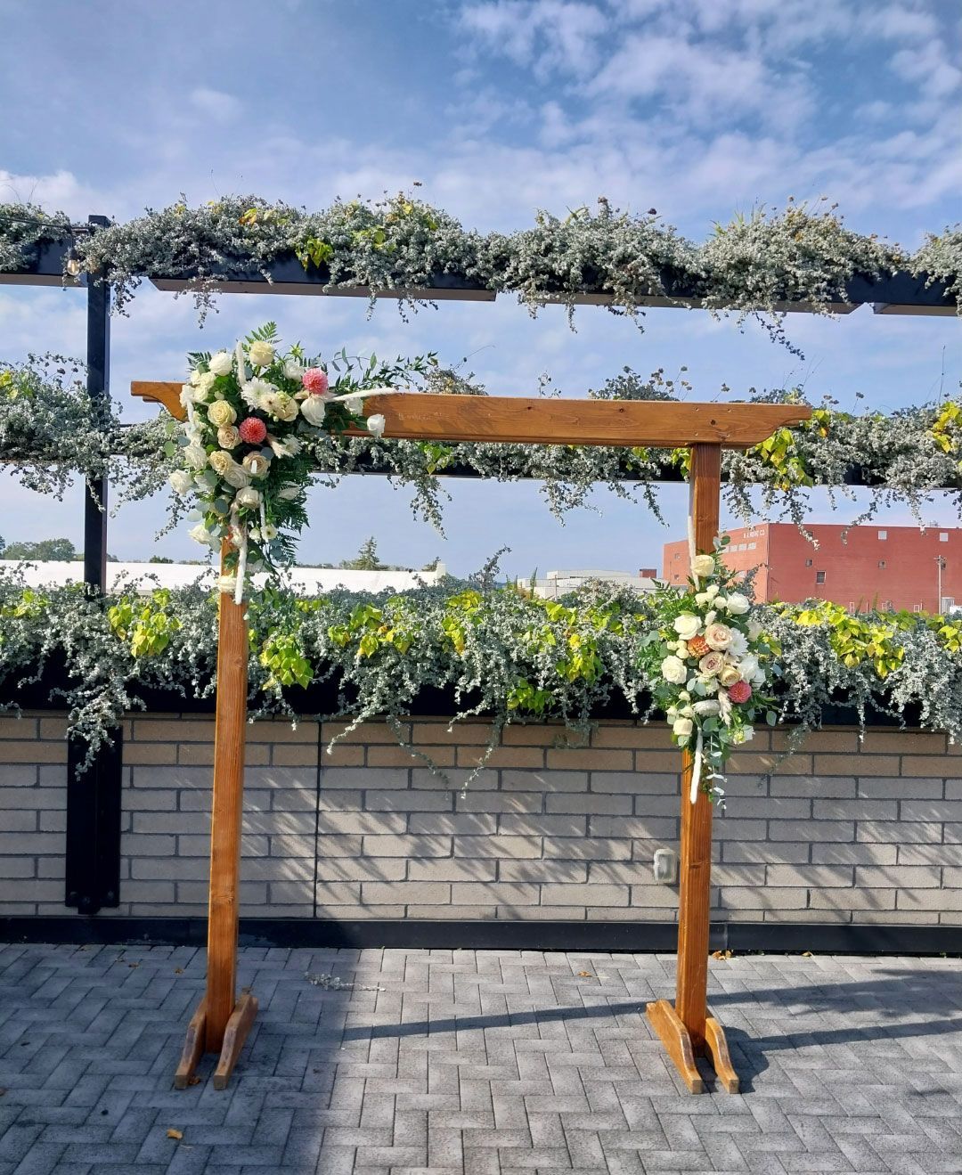 Wooden wedding arch decorated with flowers against a brick wall and a blue sky.