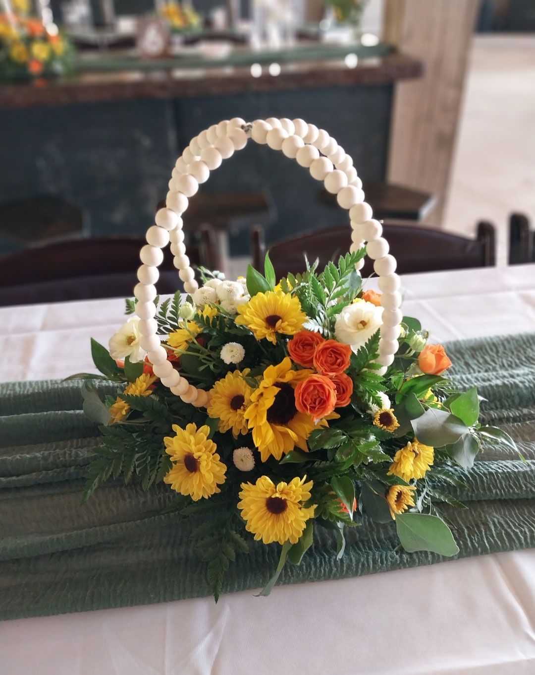 Flower arrangement in a basket with sunflowers, orange roses, and white handle on a green table runner.