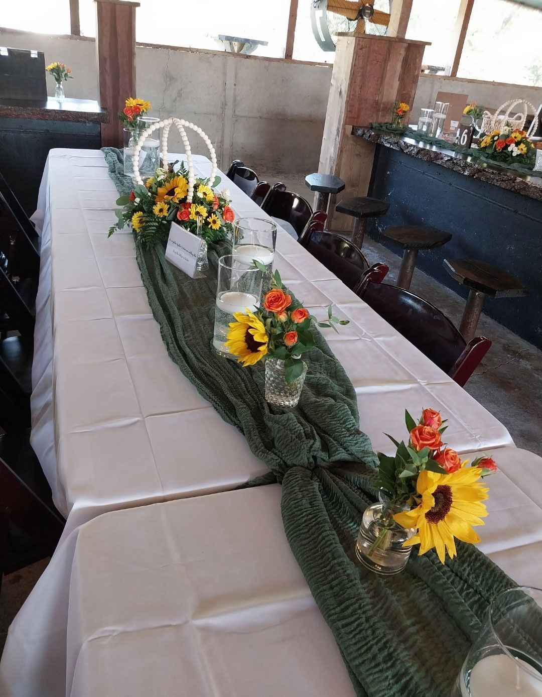 Long table set for a celebration, with white tablecloth, green runner, and flower arrangements.