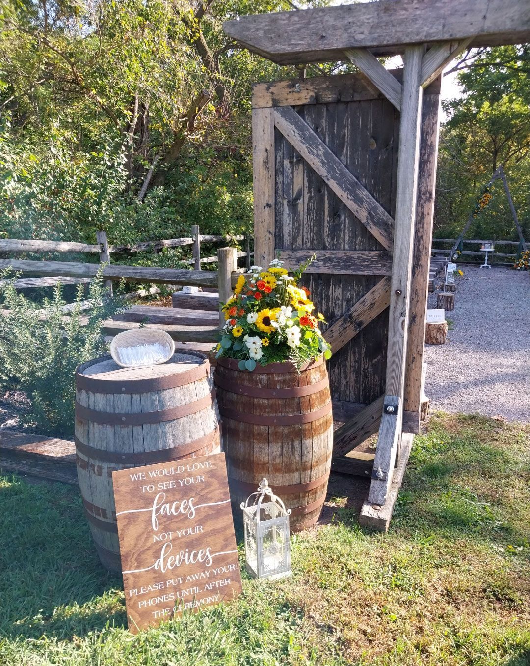 Wooden wedding decor: barrels, floral arrangement, sign, lantern, and gate against a wooded background.