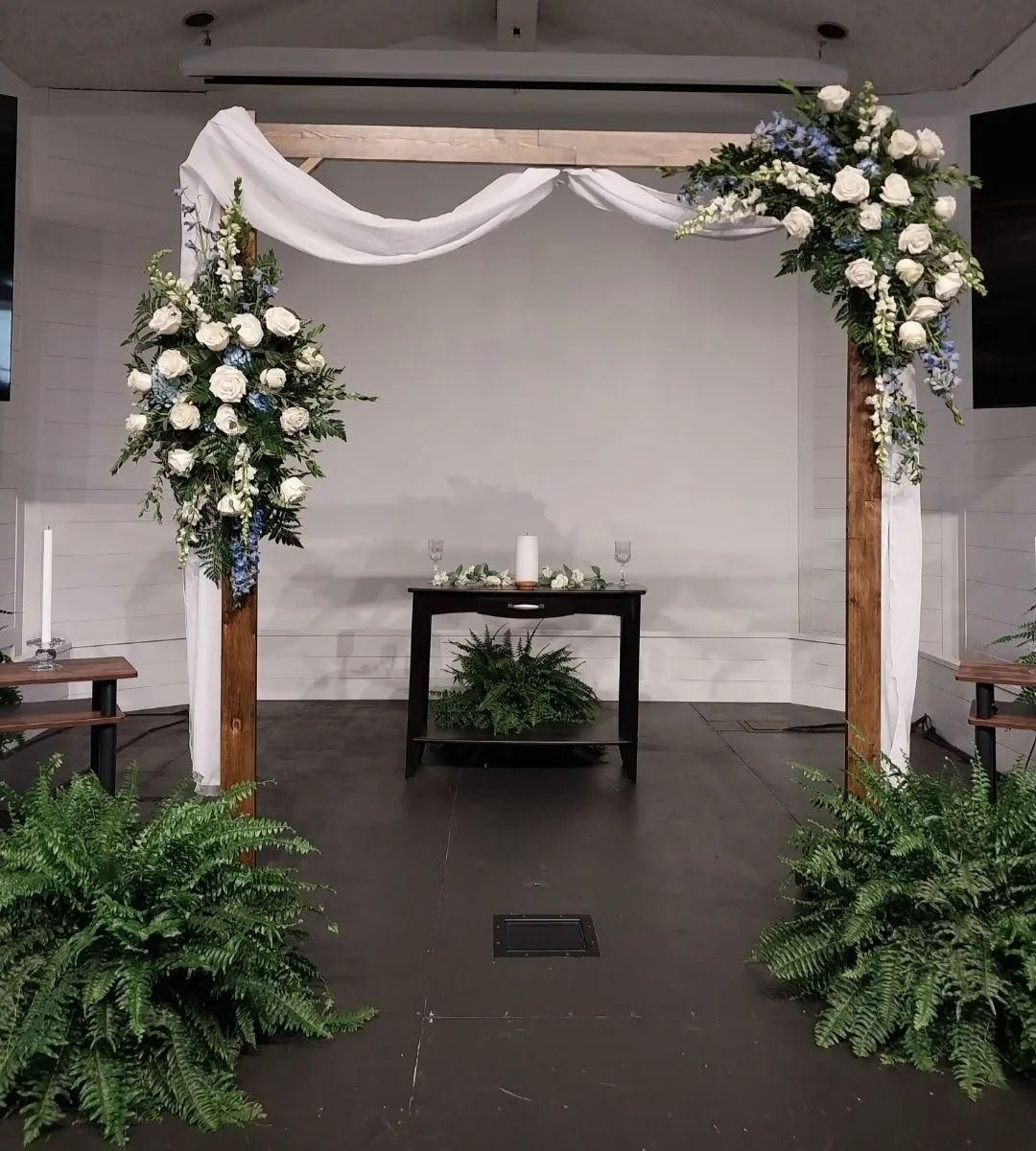 Wedding ceremony setup: wooden arch draped with fabric, decorated with white and blue flowers; ferns at base.