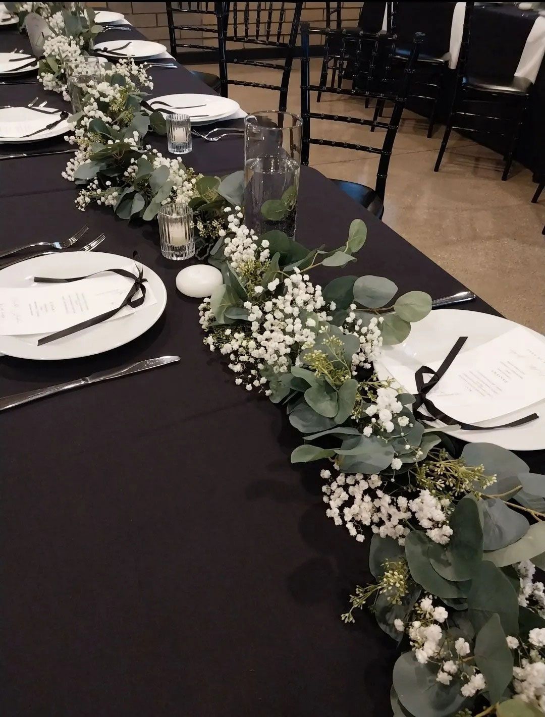 Long table set with black tablecloth, white plates, and floral garland centerpiece.