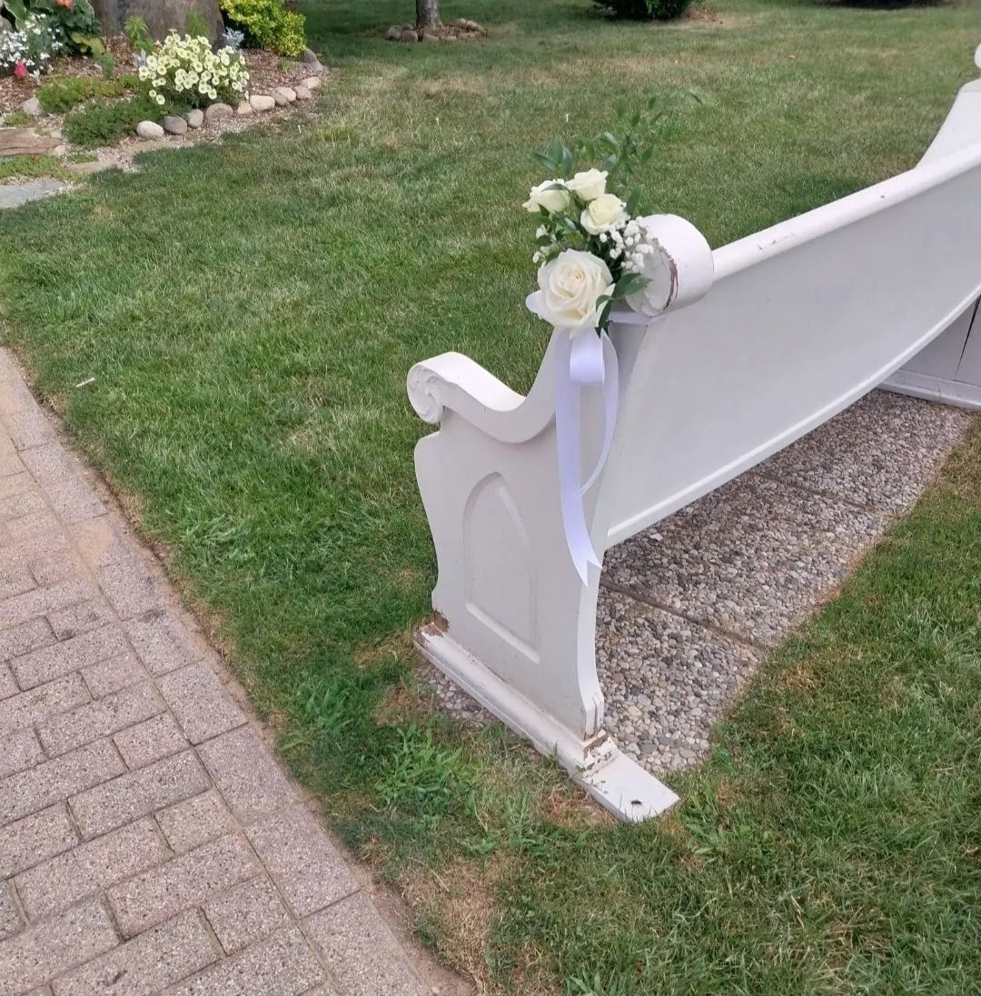 White church pew with floral arrangement and ribbon on green lawn.