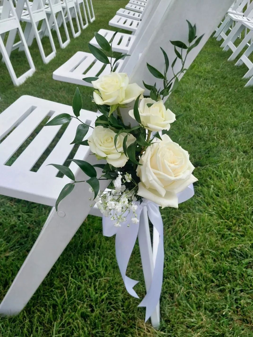 White chairs decorated with roses and greenery tied with white ribbon on grass.