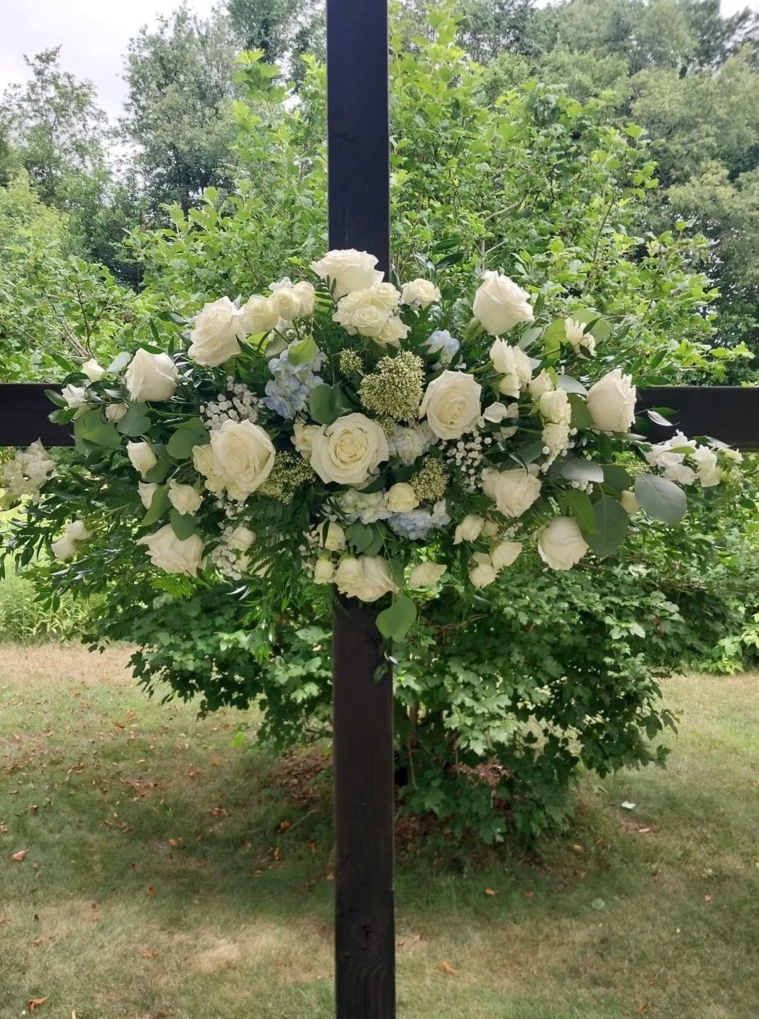White and blue floral arrangement on a black cross, set against green foliage and trees.