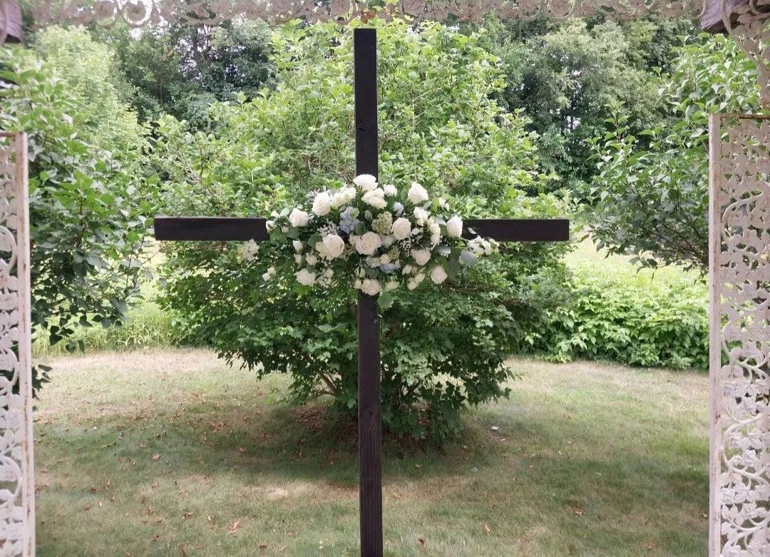 Dark wooden cross decorated with white and blue flowers, set against a backdrop of green foliage.