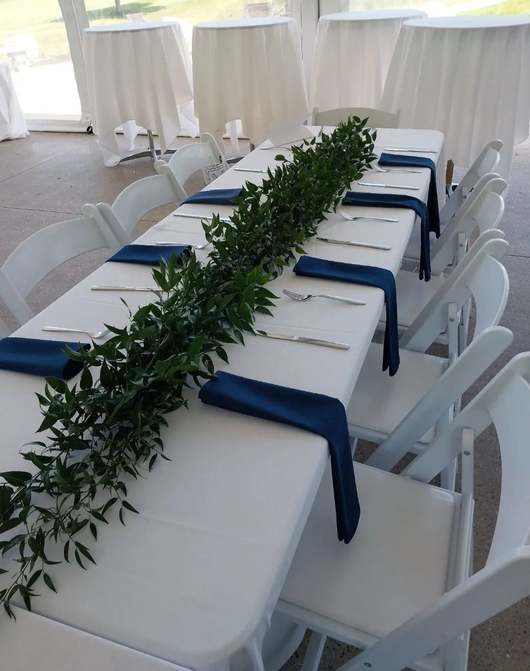 Long rectangular table set for a formal event with white linens, blue napkins, and greenery. White chairs surround the table.