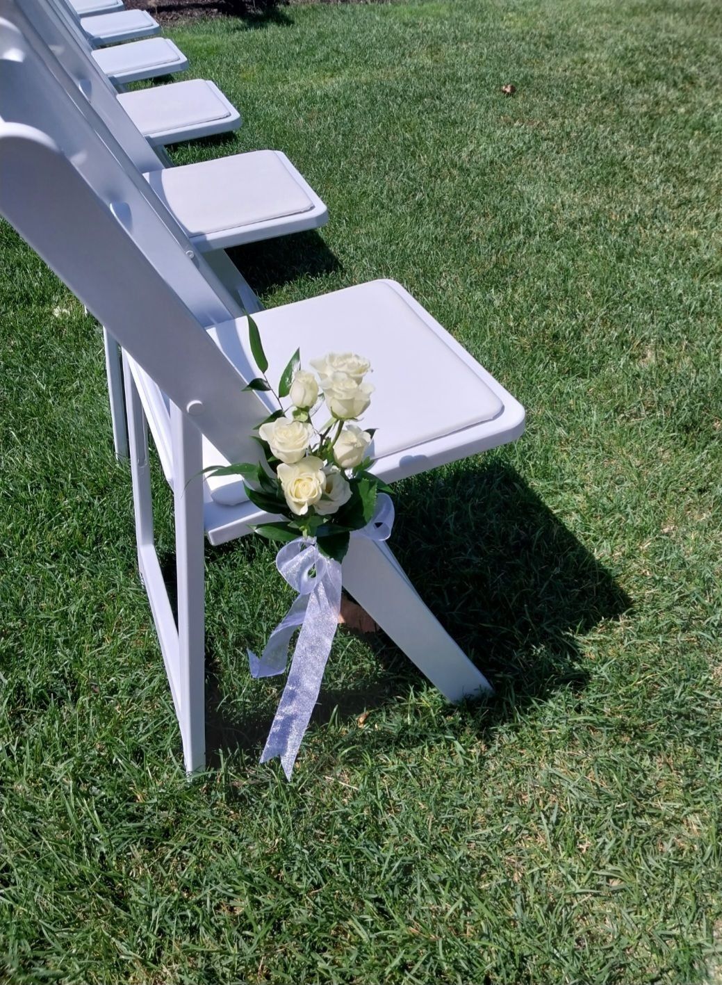 White folding chairs with floral arrangements, set up on green grass for an event.