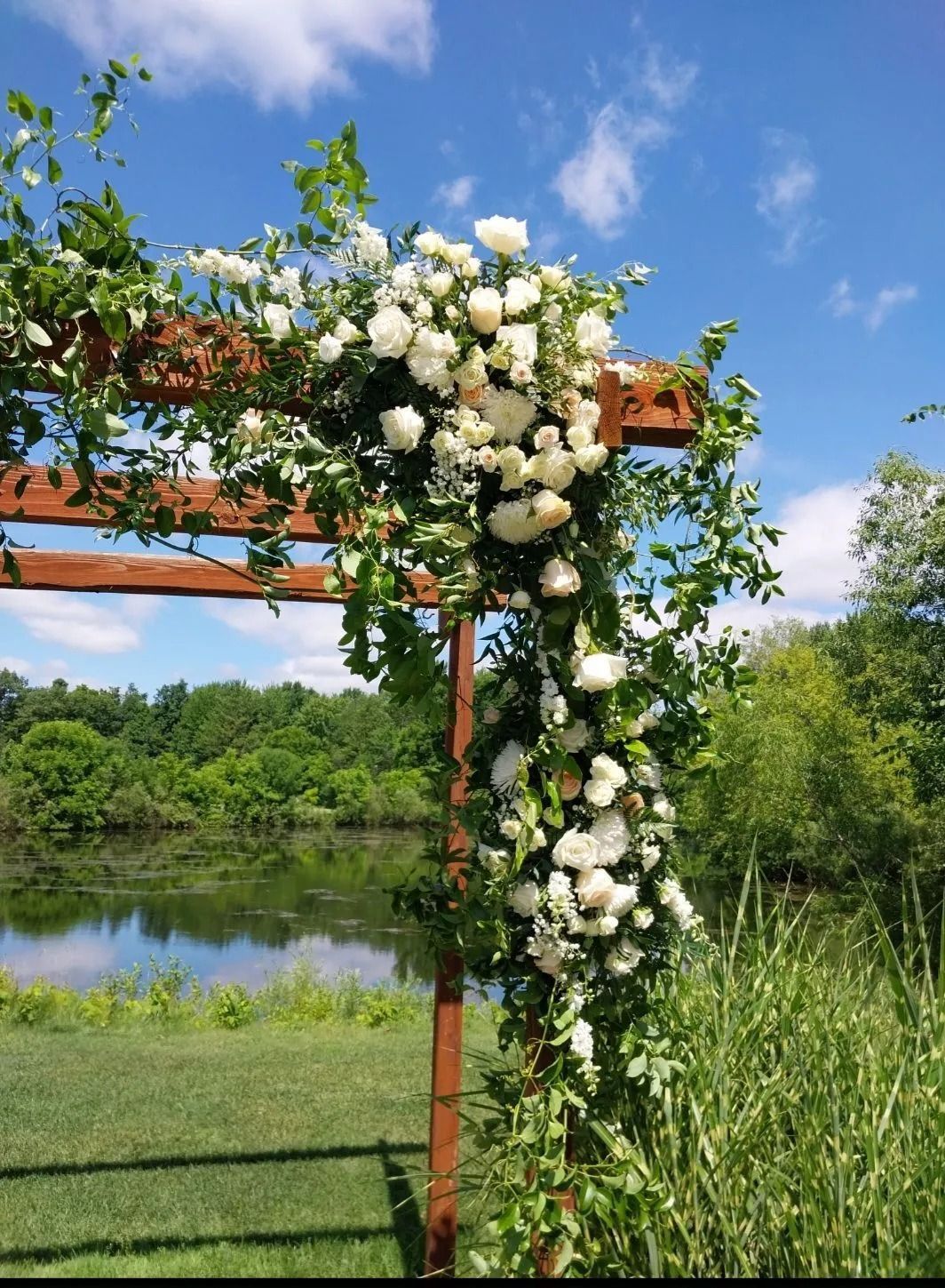 Wooden arbor decorated with white flowers and greenery, against a backdrop of lake, trees, and blue sky.