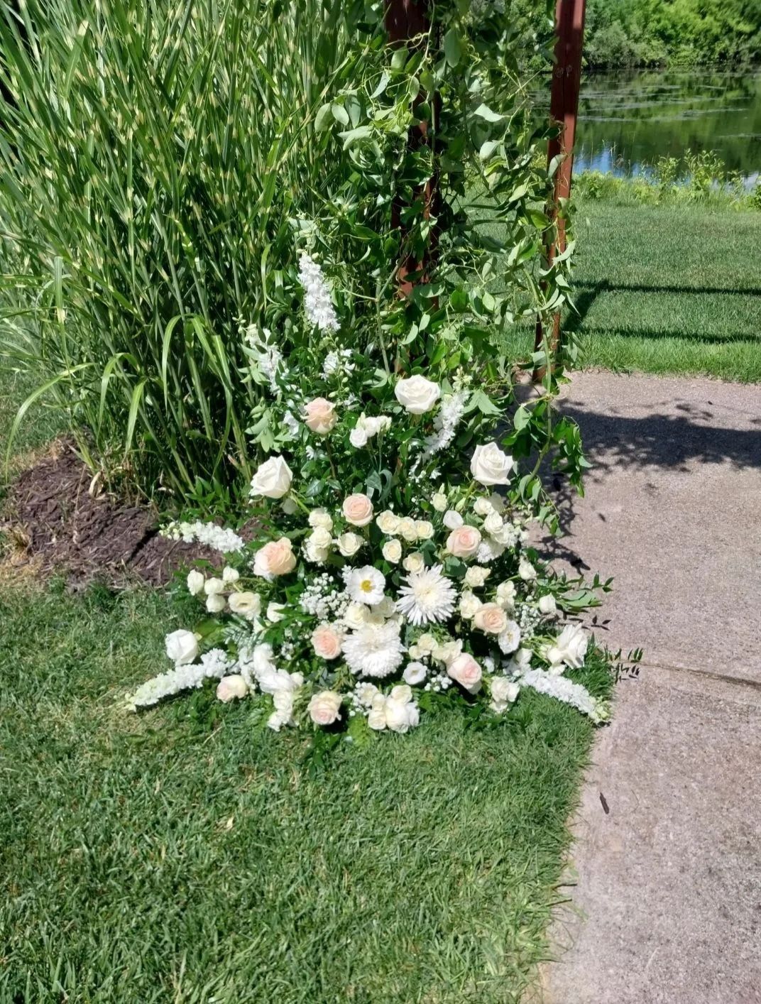 Floral arrangement of white and cream roses, daisies, and greenery on grass next to a paved path and tall foliage.